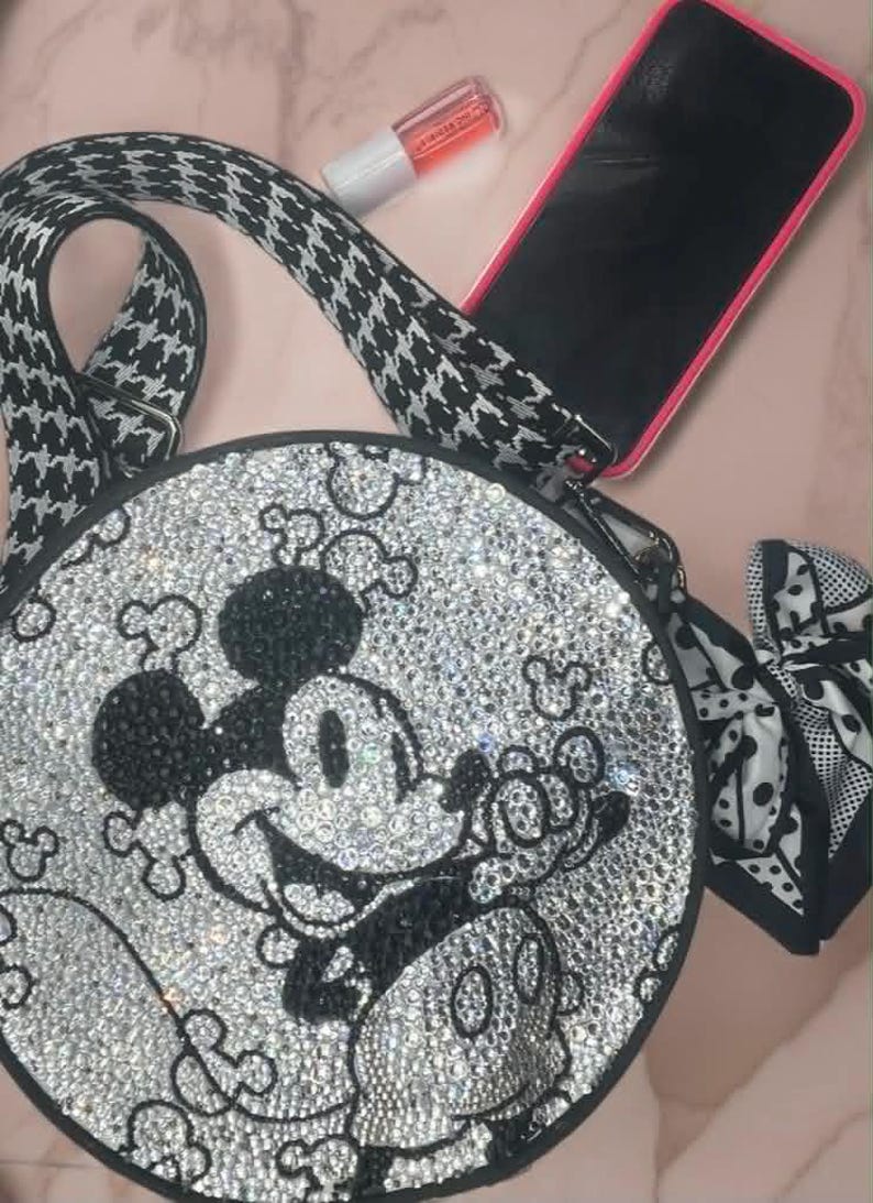 May include: A round crossbody bag featuring a crystal-encrusted Mickey Mouse design in black and clear stones. The bag has a black and white houndstooth patterned strap, a black and white polka dot side bow, a smartphone, and a small lip gloss tube.