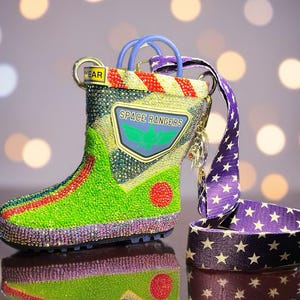 Puede incluir: Un bolso con forma de bota cubierto de pedrer&iacute;a multicolor que presenta un parche de Space Rangers con un emblema de ala verde. El bolso tiene una correa morada con estrellas blancas, detalles a rayas rojas y amarillas y una etiqueta YEAR.