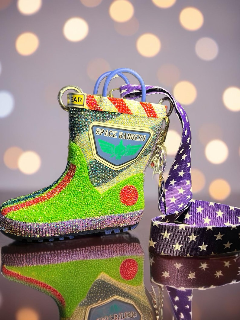 Puede incluir: Un bolso con forma de bota cubierto de pedrer&iacute;a multicolor que presenta un parche de Space Rangers con un emblema de ala verde. El bolso tiene una correa morada con estrellas blancas, detalles a rayas rojas y amarillas y una etiqueta YEAR.