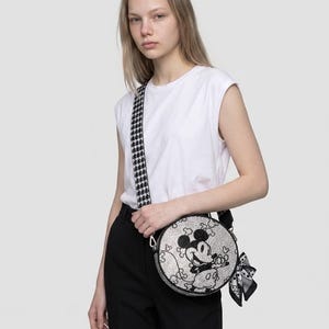 May include: A round crossbody bag featuring a black and white graphic of Mickey Mouse on the front. The bag has a textured surface, a black and white houndstooth patterned shoulder strap, and a small black and white patterned scarf tied to the side.