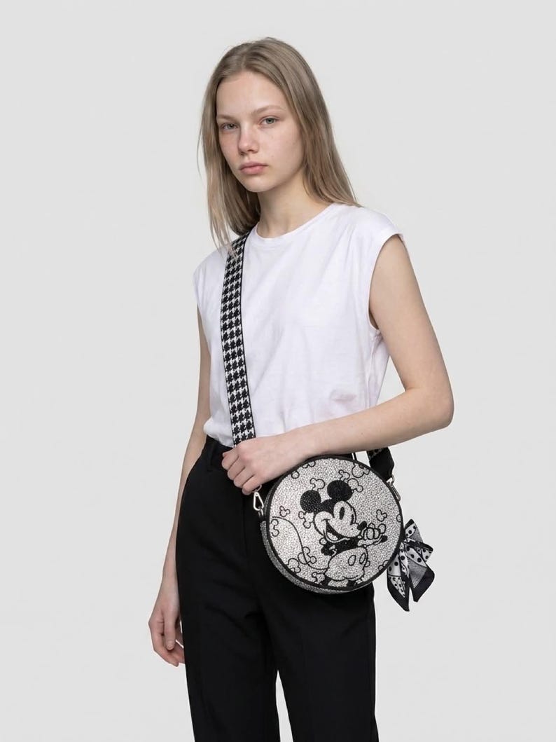 May include: A round crossbody bag featuring a black and white graphic of Mickey Mouse on the front. The bag has a textured surface, a black and white houndstooth patterned shoulder strap, and a small black and white patterned scarf tied to the side.