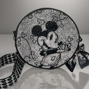 May include: A round black crossbody bag featuring a crystal-encrusted surface that depicts a Mickey Mouse character graphic. The bag includes a black and white houndstooth patterned strap and a decorative black and white patterned fabric bow.