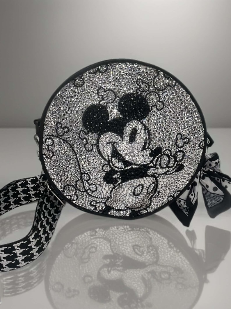 May include: A round black crossbody bag featuring a crystal-encrusted surface that depicts a Mickey Mouse character graphic. The bag includes a black and white houndstooth patterned strap and a decorative black and white patterned fabric bow.