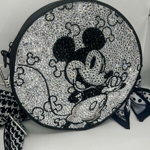 May include: A round crossbody bag covered in clear and black rhinestones forming a Mickey Mouse character design. The bag features a black border, a silver-toned metal clip, and a black and white patterned fabric strap with a matching fabric accent.