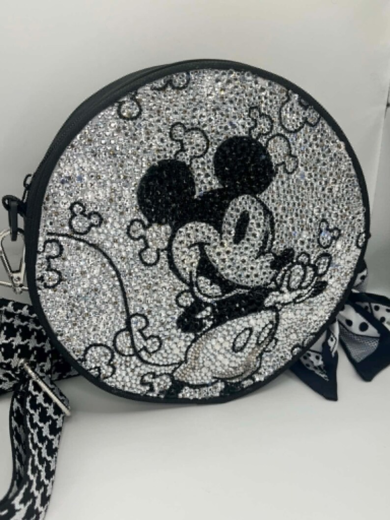May include: A round crossbody bag covered in clear and black rhinestones forming a Mickey Mouse character design. The bag features a black border, a silver-toned metal clip, and a black and white patterned fabric strap with a matching fabric accent.