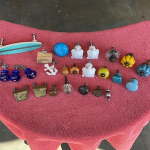 May include: A collection of various cabinet knobs and pulls in diverse shapes, colors, and materials. The assortment includes a surfboard-shaped pull, white turtle-shaped knobs, a white anchor, and several round, faceted, and metallic hardware pieces.