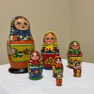 May include: A set of six hand-painted wooden nesting dolls of varying sizes. Each doll features a painted face with dark hair, a headscarf, and a colorful apron with floral details in shades of blue, orange, yellow, red, and green.