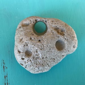 May include: A light tan, porous stone with a rough, uneven texture. The stone features one large, circular hole that passes completely through the center, along with several smaller, shallow circular indentations across its surface.
