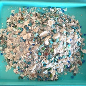 May include: A rectangular turquoise tray filled with a mixture of small white seashells, fragments of blue and green sea glass, and various small round blue beads. The collection of materials is piled in the center of the tray.