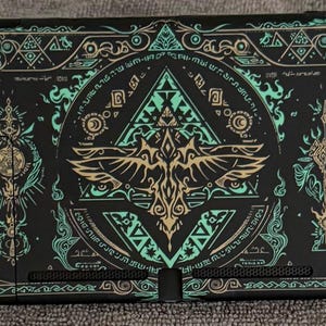以下が含まれることがあります： 黒色のNintendo Switchコンソール、カスタムデザイン。 ターコイズとゴールドの幾何学模様と、翼を広げた鳥の中央エンブレムが特徴です。 コンソールのエッジにはゴールドのアクセントがあります。