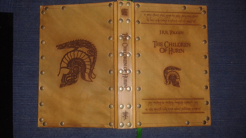 May include: A brown leather-bound book with a title embossed in gold lettering: "The Children of Hurin" by J.R.R. Tolkien. The cover features a dragon and a helmet design.