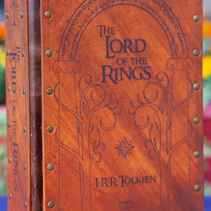 May include: A brown leather-bound book with the title "The Lord of the Rings" and the author's name "J.R.R. Tolkien" embossed on the cover. The book has a decorative design of trees and a star.