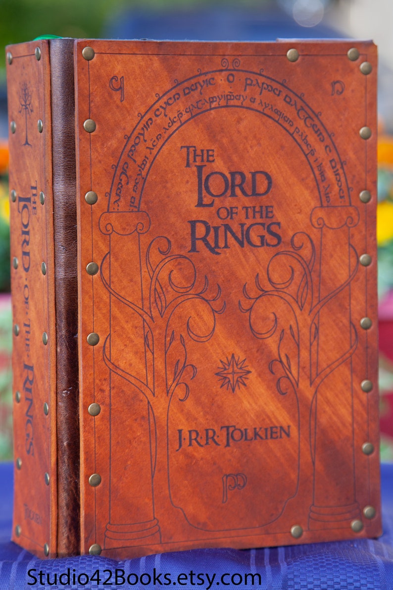 May include: A brown leather-bound book with the title "The Lord of the Rings" and the author's name "J.R.R. Tolkien" embossed on the cover. The book has a decorative design of trees and a star.
