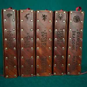 May include: Five leather-bound books with embossed titles and author names. The titles are "A Game of Thrones", "A Clash of Kings", "A Storm of Swords", "A Feast for Crows", and "A Dance with Dragons". The author is George R.R. Martin.