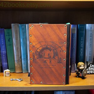 May include: A brown leather-bound journal with metal accents and a decorative design on the front cover. The design features a circular archway with a quote below it that reads "Let us not be too particular, it is better to have old secondhand happiness than none at all."