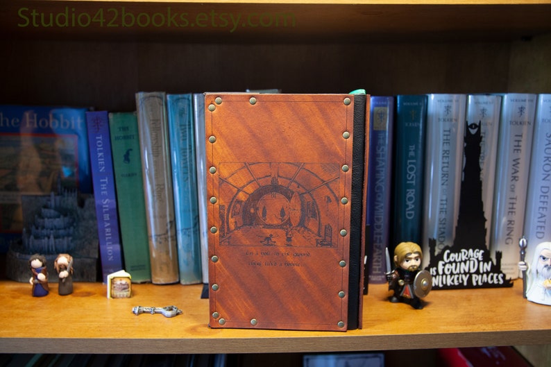 May include: A brown leather-bound journal with metal accents and a decorative design on the front cover. The design features a circular archway with a quote below it that reads "Let us not be too particular, it is better to have old secondhand happiness than none at all."