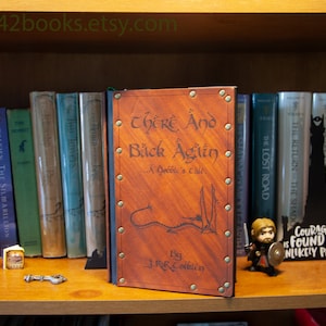 May include: A brown leather-bound book with the title "There And Back Again... A Hobbit's Tale" by J.R.R. Tolkien. The book is on a bookshelf with other books, including "The Hobbit" and "The Lord of the Rings" series.