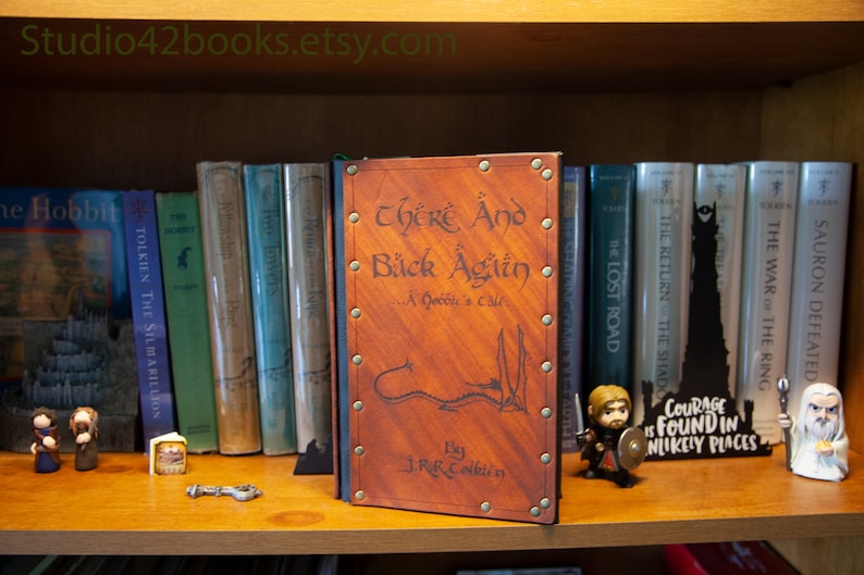 May include: A brown leather-bound book with the title "There And Back Again... A Hobbit's Tale" by J.R.R. Tolkien. The book is on a bookshelf with other books, including "The Hobbit" and "The Lord of the Rings" series.