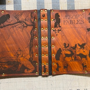 May include: A brown leather-bound book with gold accents and the title "Aesop's Fables" embossed on the cover. The cover features various animal illustrations, including a fox, a crow, a lion, and a bear.