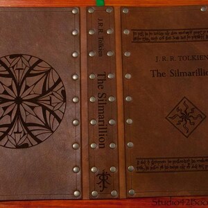 May include: A brown leather-bound book with the title "The Silmarillion" by J.R.R. Tolkien embossed on the cover. The book has decorative metal accents and a unique design on the front cover.