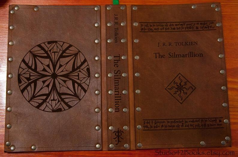 May include: A brown leather-bound book with the title "The Silmarillion" by J.R.R. Tolkien embossed on the cover. The book has decorative metal accents and a unique design on the front cover.