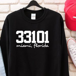 Peut inclure: Un sweat-shirt noir à manches longues et col rond avec le texte imprimé en blanc 33101 et les mots miami, florida dans une police cursive. Le vêtement est présenté sur un cintre noir devant un mur en briques blanches.