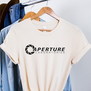 Peut inclure: T-shirt crème avec le logo Aperture Laboratories en noir. Le t-shirt est suspendu à un cintre en bois, avec une chemise en jean et un chapeau marron en arrière-plan. Le t-shirt est noué en bas.