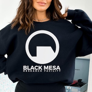Puede incluir: Sudadera negra con un logotipo blanco y el texto "BLACK MESA RESEARCH FACILITY". El logotipo es un círculo blanco con una forma blanca en su interior. La sudadera es negra.