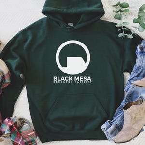 Puede incluir: Sudadera con capucha verde oscuro con un diseño gráfico blanco y el texto "BLACK MESA RESEARCH FACILITY". La imagen incluye vaqueros azules, botines color canela y una bufanda a cuadros. Un atuendo casual.