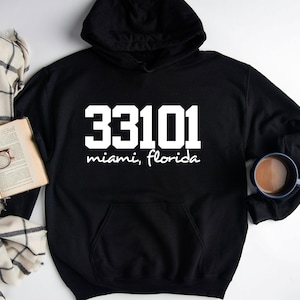 Op de afbeelding: Een zwarte hoodie met de witte tekst 33101 in een vet bloklettertype, met daaronder de woorden miami, florida in een wit handschriftlettertype. Het kledingstuk heeft een kangoeroezak aan de voorzijde en lange mouwen met geribbelde boorden.