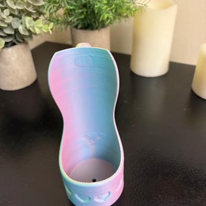 May include: A 3D-printed, pastel-colored holder with a gradient of pink, blue, and a hint of yellow. The holder has a curved shape with a heart-shaped design at the base. The object is sitting on a dark surface.