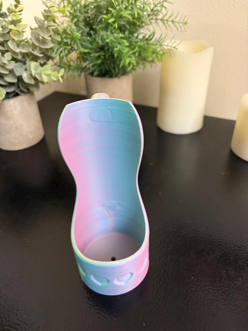 May include: A 3D-printed, pastel-colored holder with a gradient of pink, blue, and a hint of yellow. The holder has a curved shape with a heart-shaped design at the base. The object is sitting on a dark surface.
