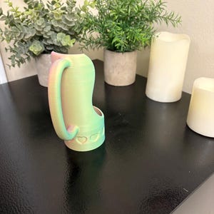 May include: A 3D-printed cat-shaped watering can with a handle and a spout. The can is gradient-colored, transitioning from green to pink. The base features heart-shaped cutouts. The can is placed on a black surface, with potted plants and candles in the background.