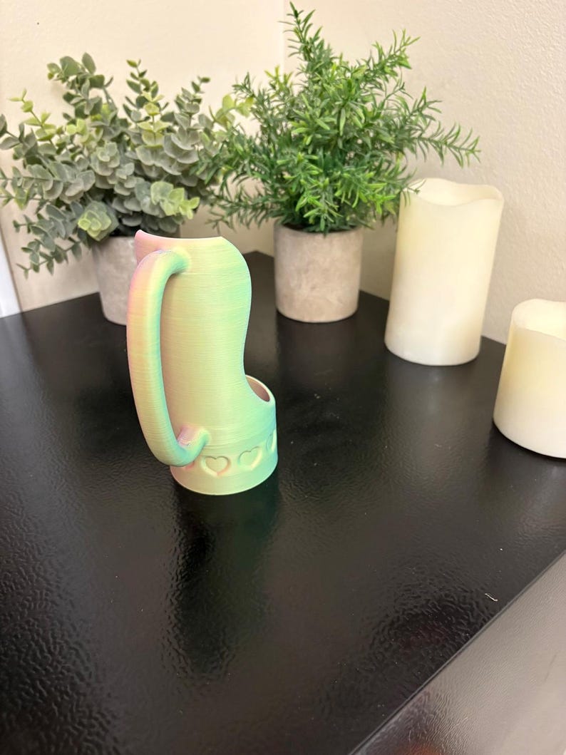 May include: A 3D-printed cat-shaped watering can with a handle and a spout. The can is gradient-colored, transitioning from green to pink. The base features heart-shaped cutouts. The can is placed on a black surface, with potted plants and candles in the background.