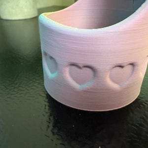 May include: A light pink, cylindrical container with heart-shaped cutouts. The container has a rounded base and a flared top. The edges are highlighted with a subtle blue-green gradient. The container is likely 3D-printed.