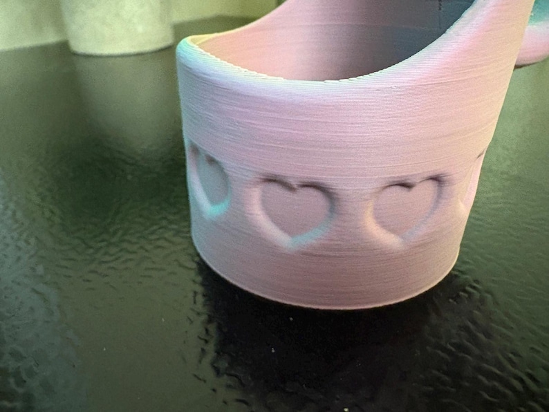 May include: A light pink, cylindrical container with heart-shaped cutouts. The container has a rounded base and a flared top. The edges are highlighted with a subtle blue-green gradient. The container is likely 3D-printed.