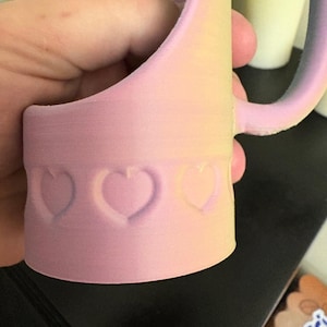 May include: A pastel pink mug with a heart-shaped design. The mug has a handle and a cylindrical base with three embossed hearts. The colors transition from pink to yellow and purple.