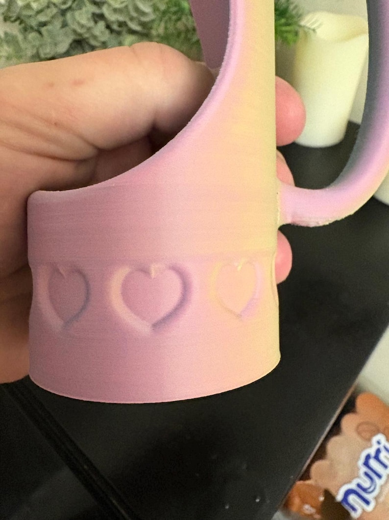 May include: A pastel pink mug with a heart-shaped design. The mug has a handle and a cylindrical base with three embossed hearts. The colors transition from pink to yellow and purple.