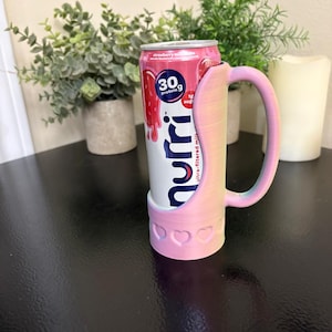 May include: A pink and white 3D-printed can holder with a handle, holding a strawberry milk drink. The holder has a heart design at the base. The drink can features the text "30g protein" and "strawberry milk".