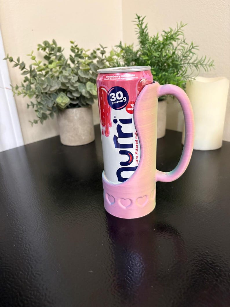 May include: A pink and white 3D-printed can holder with a handle, holding a strawberry milk drink. The holder has a heart design at the base. The drink can features the text "30g protein" and "strawberry milk".