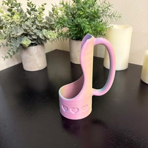 May include: A pastel-colored, 3D-printed phone holder with a handle. The holder is decorated with heart-shaped cutouts and gradients of pink, yellow, and blue. The holder is on a black surface, with potted plants and candles in the background.