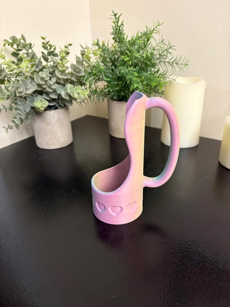 May include: A pastel-colored, 3D-printed phone holder with a handle. The holder is decorated with heart-shaped cutouts and gradients of pink, yellow, and blue. The holder is on a black surface, with potted plants and candles in the background.