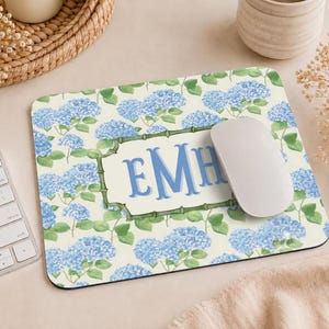 May include: A rectangular mouse pad featuring a pattern of blue hydrangea flowers and green leaves on a cream background. In the center, a bamboo-style frame encloses the blue monogram letters EMH. A white computer mouse rests on the surface.