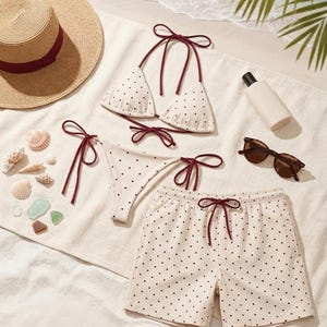 May include: A beach-themed flat lay featuring a straw sun hat with a burgundy band, a polka-dot bikini, and matching swim shorts. Sunglasses and sunscreen are also present, suggesting a day at the beach. Seashells and sea glass add to the coastal aesthetic.