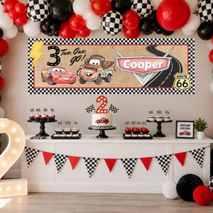May include: A birthday party display featuring a rectangular banner with a race car theme, displaying the text "Two. One. go!" and "Cooper" alongside illustrations of two cars, a Route 66 sign, and a lightning bolt, surrounded by a balloon arch.