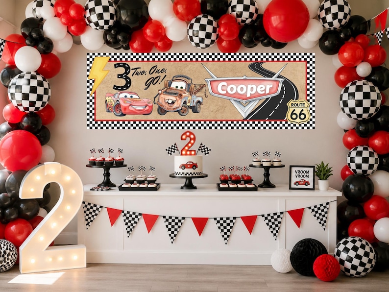 May include: A birthday party display featuring a rectangular banner with a race car theme, displaying the text "Two. One. go!" and "Cooper" alongside illustrations of two cars, a Route 66 sign, and a lightning bolt, surrounded by a balloon arch.