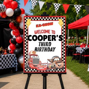 May include: A rectangular birthday sign on a black easel featuring a chequered border, yellow lightning bolts, and the text KA-CHOW WELCOME TO COOPER'S THIRD BIRTHDAY. The sign displays illustrations of a red race car and a brown tow truck.