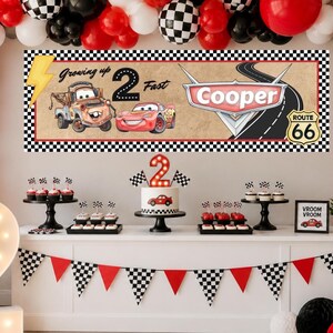 May include: A rectangular banner featuring a checkered border, a yellow lightning bolt, and the text Growing up 2 Fast. It displays cartoon cars, a road, and a Route 66 sign with the name Cooper. The banner is set against a wall with balloons.