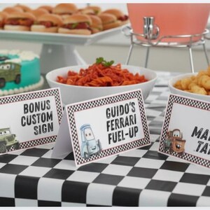 Puede incluir: Tres tarjetas de mesa rectangulares blancas con bordes de cuadros blancos y negros. Las tarjetas presentan personajes de coches de dibujos animados y el texto Bonus Custom Sign, Guido's Ferrari Fuel-Up y Tow Mater's Taters sobre un mantel.