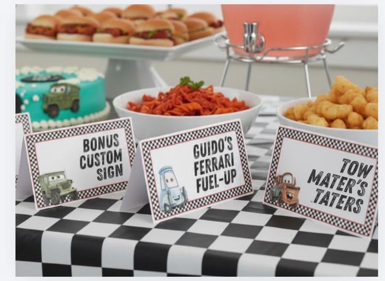 Puede incluir: Tres tarjetas de mesa rectangulares blancas con bordes de cuadros blancos y negros. Las tarjetas presentan personajes de coches de dibujos animados y el texto Bonus Custom Sign, Guido's Ferrari Fuel-Up y Tow Mater's Taters sobre un mantel.